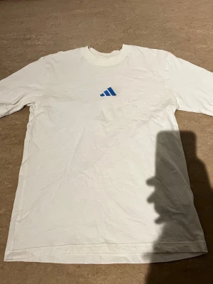 Vit Adidas t-shirt med blå logga - Säljer en vit t-shirt från Adidas med blå logga på bröstet. Klassisk rund hals och korta ärmar. Tillverkad i mjuk bomull som känns skön mot huden. Perfekt till en avslappnad stil.