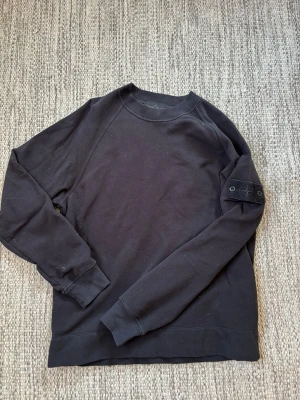 Svart sweatshirt från Stone Island - Säljer en svart sweatshirt från Stone Island med klassisk rund hals och ribbade muddar. Tröjan har den ikoniska Stone Island-loggan på ärmen och är tillverkad i mjukt bomullsmaterial. Perfekt för en clean och stilren look.