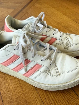 Adidas sneakers - Ett par Adidas sneakers i vitt med rosa ränder. Skorna har vita skosnören och en vit sula. Skorna har en logotyp på plösen och på sidan.  Strl 35  Hämtas i Nacka eller skickas mot porto.   
