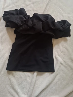 Svart offshoulder topp med volang - Säljer en svart offshoulder topp med stor volangdetalj över axlarna. Toppen har en tight passform och ärmlös design som ger en riktigt statement look. Perfekt för dig som vill sticka ut med en unik och trendig stil. Kan användas som off shoulder och med armarna uppe! Jätte fin men passade tyvärr inte mig 💕💕 be om bilder på!!