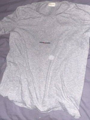 Grå Saint Laurent t-shirt - Säljer en stilren grå t-shirt från Saint Laurent med diskret logga på bröstet. T-shirten har klassisk passform, rund halsringning och är tillverkad i mjuk bomull. Perfekt för en clean och avslappnad look. Äkta tröja nypris 3000kr