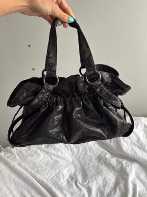 Svart handväska med metallic-look - Snygg svart handväska i glansigt material med metallic-effekt. Väskan har två rejäla handtag, rynkade detaljer och stora ringar i metall. Rymlig insida med tryckknappsstängning och cool, lite slouchy form. Perfekt för dig som gillar statement-accessoarer.