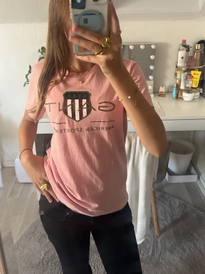 Rosa GANT t-shirt med tryck - Säljer en rosa t-shirt från GANT med stort tryck och logga framtill. Klassisk rund hals och korta ärmar. Skön och avslappnad passform, perfekt till jeans eller shorts. Tillverkad i mjuk bomull för en bekväm känsla. Det är stolek 13-14år men den passar mig som snart är 15år och nästan 170cm:)