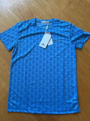 Blå mönstrad t-shirt från Lacoste - Snygg blå t-shirt från Lacoste med geometriskt mönster över hela plagget. Klassisk rund halsringning och korta ärmar. Tillverkad i mjukt material som känns skönt mot huden. Perfekt för dig som vill ha en stilren men ändå unik look.