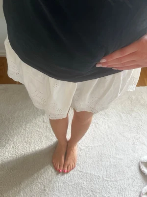 Vit kort kjol med broderade detaljer - Söt vit kort kjol med broderade hålmönster och elastisk midja. Kjolen har en lätt volangkant nedtill och är perfekt för varma dagar. Tillverkad i ett tunt och luftigt material som känns skönt mot huden. Köpt i USA