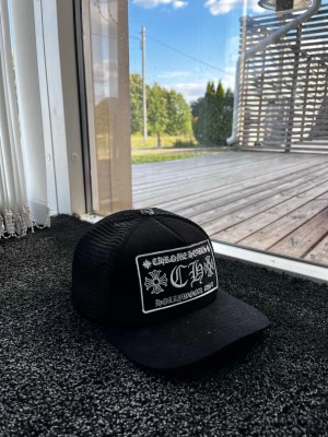 Svart truckerkeps med tryck - Snygg svart truckerkeps med vit brodyr framtill och mesh på sidorna och bak. Kepsen har en böjd skärm och justerbar snapback. Framtill står det 'CH' och dekoreras av blommor och text. Perfekt för en streetwear-look. Skick 10/10. Pris går att diskutera✅