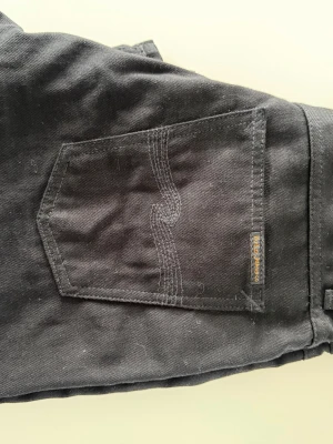 Svarta jeans från Nudie Jeans w 30 - Säljer ett par svarta jeans från Nudie Jeans med klassisk femficksmodell och raka ben. Jeansen har snygga kontrastsömmar på bakfickan och silverfärgade nitar. Perfekta för dig som gillar en clean och stilren look.