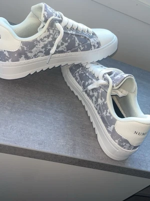 Vita och grå cloud  NUMERIS - BOX INGÅR Snygga sneakers från NUMERIS i vitt och grått camomönster. Skorna har tjock vit sula med räfflad kant, vita skosnören och detaljer i vitt konstläder. Perfekta för dig som vill sticka ut med en unik och modern look.