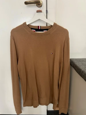 Beige stickad tröja Tommy Hilfiger - Snygg beige stickad tröja från Tommy Hilfiger i Pima cotton blend med cashmere. Tröjan har rund halsringning, långa ärmar och en liten broderad logga på bröstet. Perfekt för en stilren och avslappnad look.
