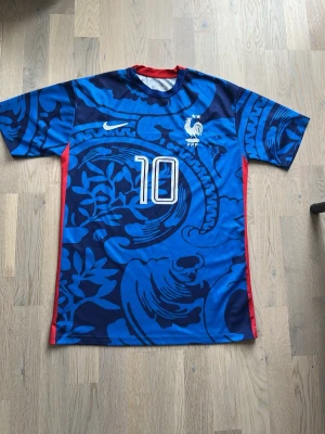 Blå Frankrike fotbollströja Mbappé - Snygg blå fotbollströja från Nike med Frankrikes landslagsmärke och nummer 10 på bröstet. Baksidan har Mbappé och nummer 10 tryckt. Tröjan har ett unikt mönster i olika blå nyanser och röda detaljer på sidorna. Perfekt för dig som älskar fotboll och vill sticka ut.