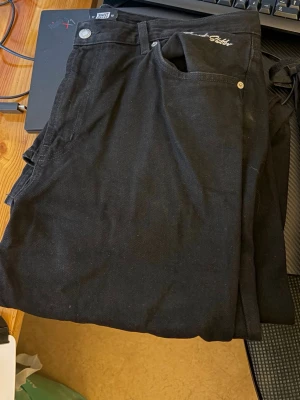 Svarta raka jeans med broderad detalj - Säljer ett par svarta jeans med rak passform och fem klassiska fickor. Jeansen har en broderad textdetalj vid fickan och silverfärgade nitar. Materialet ser ut att vara bomull med lite stretch. Perfekta för en clean och stilren look.