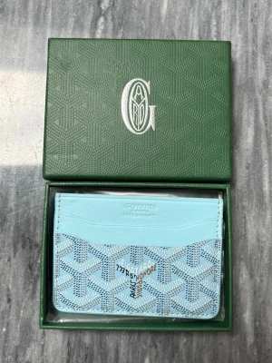 Ljusblå korthållare från Goyard - Snygg ljusblå korthållare från Goyard med klassiskt mönster och flera kortfack. Tillverkad i läder och canvas med tryckt logotyp och adressdetaljer på framsidan. Perfekt för dig som vill ha en stilren och exklusiv accessoar.