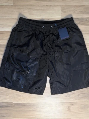 Svarta Louis Vuitton shorts - Snygga svarta shorts från Louis Vuitton med diskret monogrammönster i tyget. Shortsen har dragsko i midjan och är tillverkade i ett glansigt, lätt material som känns sportigt och exklusivt. Perfekta för dig som gillar designerplagg med subtila detaljer.