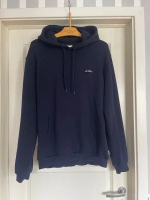 Mörkblå hoodie Les duex  - Mörkblå hoodie från Les duex med klassisk huva och dragsnören. Tröjan har en stor magficka framtill och en liten logga på bröstet. Tillverkad i mjukt material som känns skönt mot huden. Perfekt för en avslappnad och stilren look. OBS mudden har vrängt sig lite men inget som syns med blotta ögat 