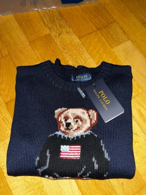 Mörkblå stickad tröja med björn - Mörkblå stickad tröja från Ralph Lauren med ikonisk björn på bröstet, klädd i jeans och svart tröja med amerikansk flagga. RL-broderi i rött nedtill. Rund halsringning och ribbade muddar. Perfekt för dig som gillar klassisk preppy stil.