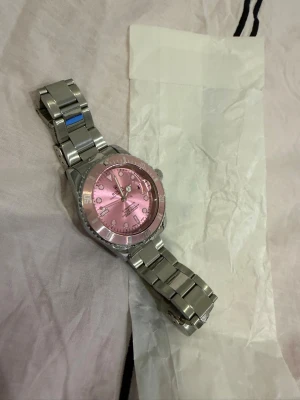 klocka  med rosa urtavla - Stilren klocka som är en  Submariner med metallarmband och en unik rosa urtavla. Klockan har tydliga indexmarkeringar, datumfönster och vridbar bezel i matchande rosa. Perfekt för dig som vill sticka ut med en klassisk men färgstark accessoar. Mvh ItsForfits 