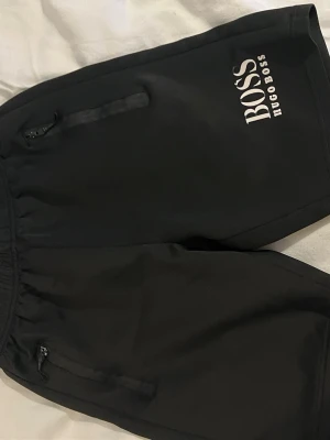 Svarta shorts från Hugo Boss - Svarta shorts från Hugo Boss med vit logga på benet. De har elastisk midja och dragkedjeförsedda fickor. Perfekta för en avslappnad och sportig stil. Materialet känns mjukt och bekvämt, troligen polyester.