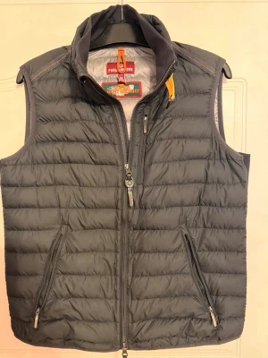 Svart dunväst från Parajumpers - Snygg svart dunväst från Parajumpers i superlätt material. Västen har hög krage, två dragkedjeförsedda fickor fram och en gul detalj vid halsen. Perfekt för lager-på-lager och enkel att matcha med olika outfits.