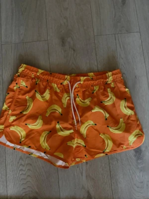 Orangea badbyxor med bananmönster - Säljer ett par färgglada orangea badbyxor med ett lekfullt bananmönster. De har resår i midjan och vit dragsko för justerbar passform. Perfekta för stranden eller poolen och tillverkade i snabbtorkande material. Stora i midjan men korta i längden 