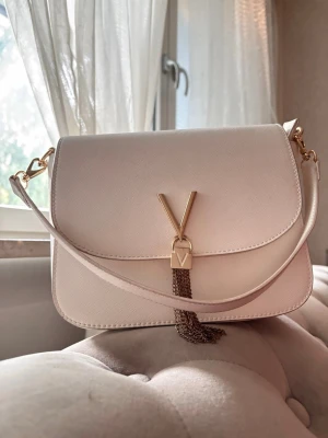 Vit Taupe axelväska från Valentino Bags - Snygg vit axelväska i taupe från Valentino Bags med stilren design. Väskan är använd ett fåtal gånger och väldigt sparsamt, så den är i princip som ny. Säljer den då jag fick den av mitt ex och därför vill bli av med den, annars är det en jättefin väska. 550 kr är mitt pris. På bild 3 finns väskans originalpris.