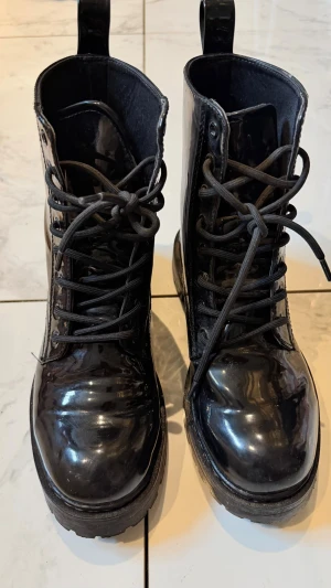 Svarta lackade snörkängor - Säljer ett par svarta kängor i glansigt lackmaterial med grov sula och snörning framtill. Kängorna har dragflikar bak för enkel påtagning och en chunky look som passar till många stilar.