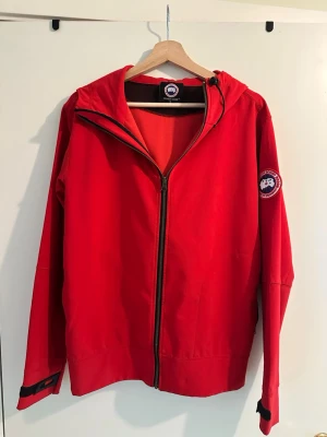 Röd Canada Goose Vindjacka - Snygg röd Canada Goose vindjacka med huva och hel dragkedja framtill. Jackan har ett emblem på ärmen och justerbara muddar med kardborre. Perfekt för blåsiga dagar och enkel att matcha med olika outfits.