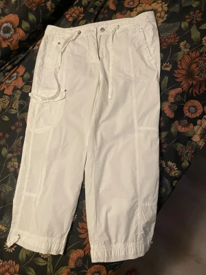 Vita cargobyxor med dragsko, Hummel  - Säljer ett par vita cargobyxor med dragsko i midjan och vid benslut. Byxorna har flera fickor, både fram och bak, samt detaljer i form av sömmar och nitar. Perfekta för en avslappnad och trendig stil. Materialet känns lätt och luftigt.