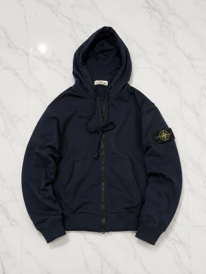 Marinblå hoodie från Stone Island - Snygg marinblå hoodie från Stone Island med dragkedja och klassisk logotyp-patch på ärmen. Huva med dragsko och mjukt material som känns skönt mot huden. Perfekt för en avslappnad streetwear-look.