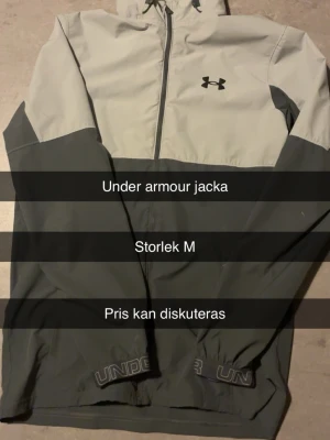 Grå vindjacka från Under Armour - Snygg tvåfärgad vindjacka från Under Armour i ljusgrått och mörkgrått. Jackan har huva, dragkedja framtill och elastiska muddar. Perfekt för blåsiga dagar och sportiga outfits. Under Armour-logga på bröstet och tryck vid midjan.