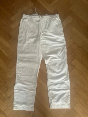Vita linnebyxor med resårmidja - Säljer ett par vita linnebyxor med avslappnad passform och resår i midjan samt dragsko. Byxorna har raka ben och en bakficka. Perfekta för en chill och stilren look under varma dagar.