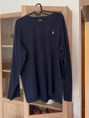 Mörkblå långärmad tröja  från Polo - En stilren mörkblå långärmad tröja från Polo Ralph Lauren med rund halsringning och diskret logga broderad på bröstet. Tillverkad i mjuk bomull som känns skön mot huden. Perfekt för en avslappnad och klassisk look.