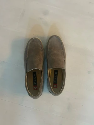 Bruna mockaloafers från Zeus - Säljer ett par gråa loafers från Zeus i mocka med klassisk slip-on design. Skorna har en diskret söm längs ovansidan och en räfflad gummisula för extra grepp. Perfekta för en avslappnad men stilren look. Storlek 42 men passar 43