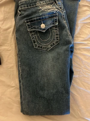Blå jeans från True Religion - Säljer ett par blå jeans från True Religion med vita kontrastsömmar och snygga broderier på bakfickorna samt en vingdetalj ovanför fickorna. Klassisk femficksmodell i denim med knappstängning. Dom har knappt används här bara lite slitage i byxbenen💕