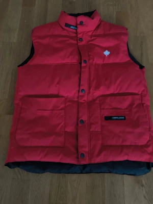 Röd väst från Canada Goose - Snygg röd dunväst från Canada Goose med hög krage, stora framfickor och tryckknappar som stängning. Västen har ett ikonisk vitt löv på och själva västen passar all väder året runt. Är inte säker på om västen är äkta men ska då vara köpt från skistarshop och fakes tillverkas inte om det är en sådan gammal modell. Tog precis ner priset från 650 till 499 då en knapp saknas