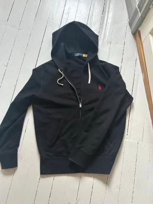Svart hoodie från Polo Ralph Lauren - Svart hoodie från Polo Ralph Lauren med dragkedja framtill, vit dragsko i huvan och röd broderad logga på bröstet. Klassisk känguruficka och ribbade muddar. Tillverkad i mjukt bomullsmaterial för en skön och avslappnad stil.