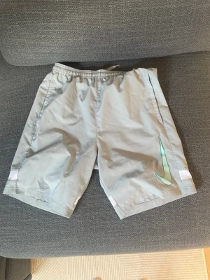 Ljusgrå Nike shorts med logga - Snygga ljusgrå shorts från Nike med elastisk midja och en stor turkos Nike-logga på ena benet. Shortsen har en sportig look och är tillverkade i ett lätt och bekvämt syntetmaterial, perfekta för träning eller chill dagar.