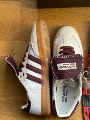Adidas x Wales Bonner sneakers vita/vinröda - Snygga Adidas x Wales Bonner sneakers i vitt och vinrött med klassiska tre ränder på sidan, grov gummisula och detaljerad plös med Wales Bonner-logga. Materialmix av läder och mocka, samt matchande vinröda skosnören. Kommer med originalkartong och dustbag.