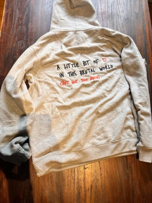 Grå hoodie med text på ryggen - Säljer en ljusgrå hoodie ifrån Mira Paris med huva och känguruficka. På ryggen står det 'A little bit of ♥ in this brutal world (but not too much)' i svart och rött. Hoodien har dragsko i huvan och ribbade muddar vid ärmar och nederkant. Perfekt för en chill och avslappnad stil.