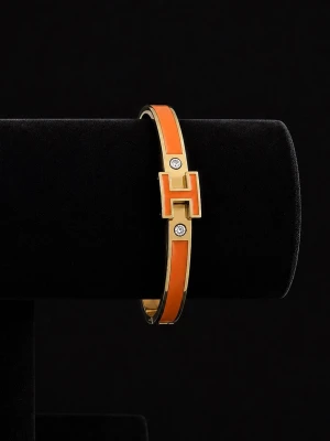 Snyggt Armabnd med H-detalj - Snyggt armband i orange och guld med en tydlig H-detalj framtill och två gnistrande stenar. Armbandet har en stilren, smal form och är tillverkat i metall med emaljerad yta. Perfekt för dig som vill ha en färgklick till din outfit.