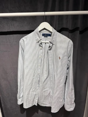Ralph Lauren skjorta - Säljer en slim fit skjorta från Ralph Lauren i vitt och blått med tunna vertikala ränder. Klassisk button-down krage, broderad logga på bröstet och knappar framtill. Tillverkad i mjuk bomull, perfekt för en clean och stilren look. Skjortan är i riktigt bra skick. Storlek S men kan passa M. Piset kan diskuteras, Skriv vid minsta fundering!