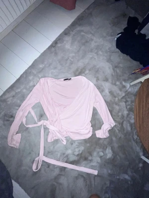 Ljusrosa omlottopp med knytband - Säljer en ljusrosa omlottopp i mjukt material med långa ärmar och knytband i midjan. Toppen har en v-ringad skärning och ger en feminin och trendig look. Perfekt att styla med jeans eller kjol för en snygg outfit.