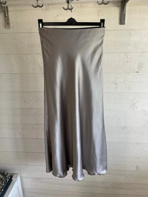 Silverfärgad långkjol i satin - Säljer en snygg silverfärgad långkjol i glansigt satinmaterial. Kjolen har en enkel och stilren design med lätt utsvängd form som ger ett elegant fall. Perfekt för dig som vill ha en trendig och tidlös look.
