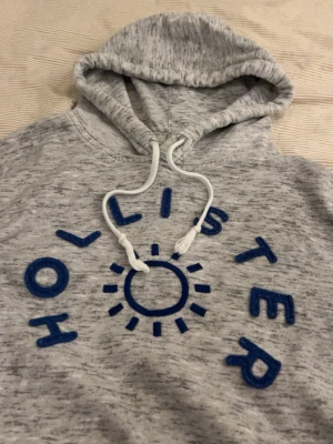 Grå hoodie från Hollister med tryck - Snygg gråmelerad hoodie från Hollister med blått broderat tryck och solmotiv på bröstet. Mjuk insida och klassisk huva med vita dragsnören. Perfekt för chill dagar och enkel att matcha med jeans eller joggers.