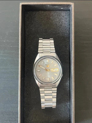 Seiko 5 Automatic – Vintage Herrklocka - Snygg vintage Seiko 5 automatisk herrklocka i mycket fint skick.  Klockan fungerar som den ska och går bra. Den har en stilren silverfärgad urtavla med guldfärgade detaljer, dag- och datumvisning samt en klassisk rostfri stållänk.  **Detaljer:**  * Seiko 5 Automatic * Automatisk mekanisk urverk * Dag- och datumvisning * Silverfärgad urtavla med guldfärgade detaljer * Rostfri stållänk * Fungerar utan problem  Säljes då den inte kommer till användning. Bättre för smalare handleder