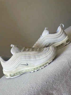 Nike Air Max 97 vita sneakers - Säljer ett par fräscha Nike Air Max 97 sneakers i vitt med ikoniska vågiga paneler och synlig Air-sula. Skorna har mesh- och läderdetaljer, silverfärgad Swoosh och reflekterande inslag. Perfekta för dig som gillar klassisk streetwear och vill ha en clean look.