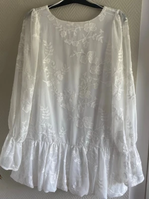 Vit blommig kortklänning med volang - Super fin vit kortklänning med blommigt mönster, transparenta långärmar och volangkant nedtill. Klänningen har rund halsringning fram och djup rygg med knytband. Perfekt för sommarens alla tillfällen.