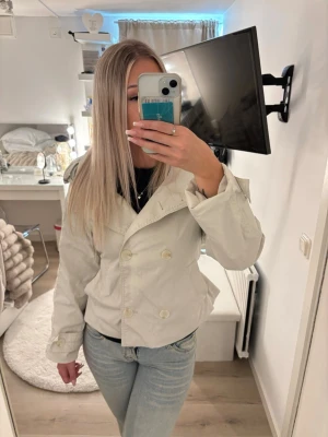 Kort beige trenchcoat med knytskärp - Snygg kort beige trenchcoat med dubbelknäppning och knytskärp i midjan. Jackan har klassisk krage, axelklaffar och är tillverkad i ett lätt tyg som passar perfekt till våren. Stilsäker och enkel att matcha till olika outfits. Nypris är 800kr och säljer pågrund av att den ej kommer till användning 