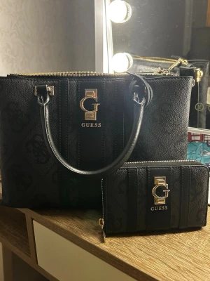 Svart handväska och plånbok Guess - Snygg svart handväska från Guess i konstläder med matchande plånbok. Väskan har guldfärgade detaljer, dubbla handtag och dragkedja. Guess-logga framtill och diskret mönster i materialet. Insidan är ljus med Guess-tryck och flera fack för bra organisering.