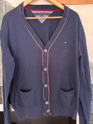 Marinblå cardigan Tommy Hilfiger - Stilren marinblå cardigan från Tommy Hilfiger med knappar framtill, två fickor och tunna röda och vita detaljer längs kanterna. Klassisk V-ringning och ribbade muddar. Perfekt för lager på lager och enkel att matcha.