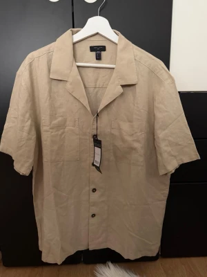 Beige linneskjorta från New Look - Säljer en beige kortärmad skjorta från New Look i luftig linneblandning. Skjortan har klassisk krage, knappar framtill och två bröstfickor. Perfekt för varma dagar och enkel att styla till olika outfits💕💗